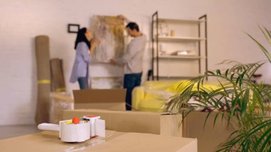 scotch tape on carton boxes near blurred couple hanging painting in new home clipart