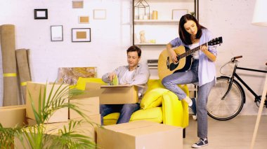 young woman playing guitar near boyfriend unpacking box in new home clipart