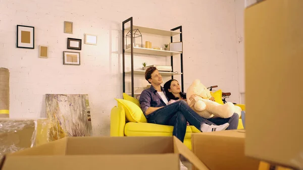 新家的一对快乐的夫妇在沙发上和泰迪熊一起在箱子边寒战 — 图库照片