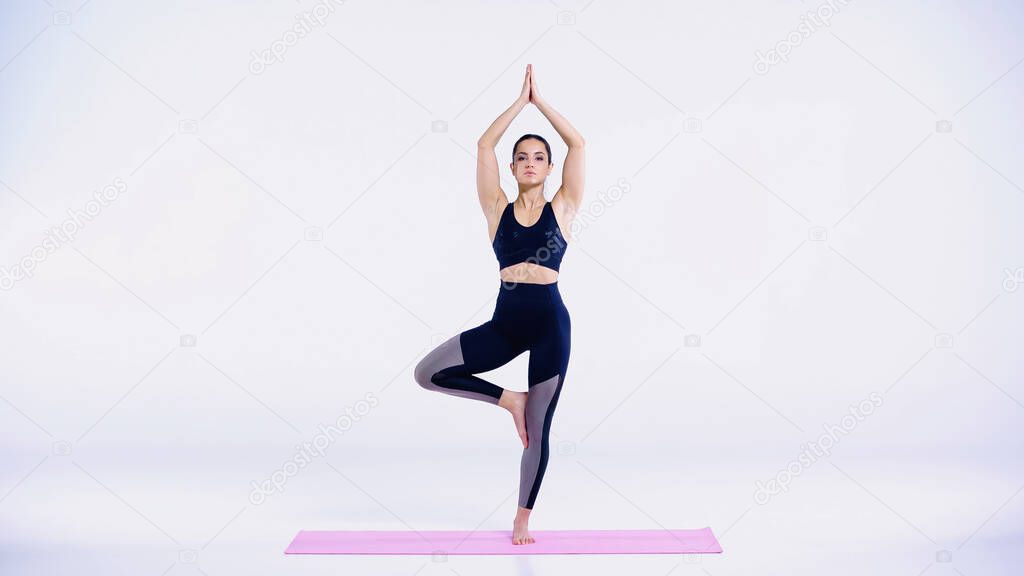 full length of young woman in sportswear standing in tree pose on yoga mat on white