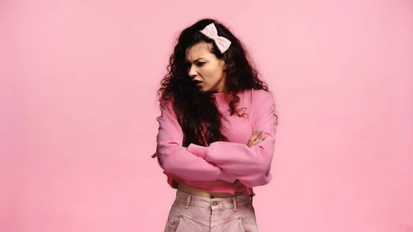 offended woman turning away while standing with crossed arms isolated on pink