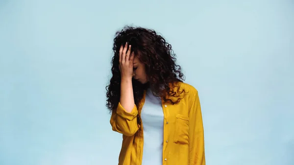 depressed woman with wavy hair covering face with hand isolated on blue