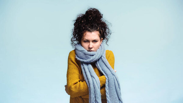 woman in warm sweater and scarf hugging herself while freezing isolated on blue