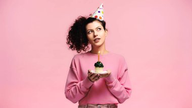 dreamy woman making wish while holding birthday cupcake isolated on pink clipart