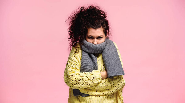 frozen woman in warm scarf and knitted sweater standing with crossed arms isolated on pink