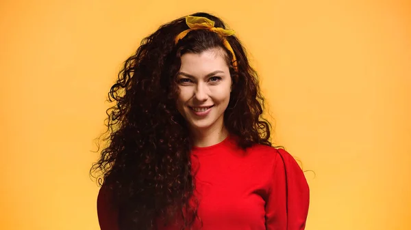 happy brunette woman with wavy hair smiling at camera isolated on orange