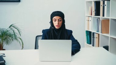 Çağrı merkezinde laptop kullanan, kulaklık takan Müslüman bir iş kadını. 