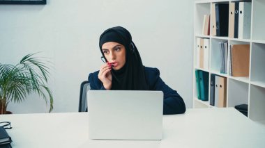 Çağrı merkezinde, bilgisayarın yanında mikrofon kullanan, kulaklıklı Müslüman bir iş kadını. 