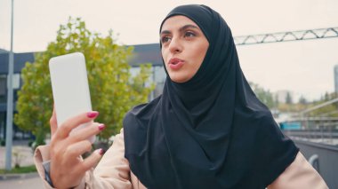 muslim woman in hijab talking while having video call on urban street  clipart