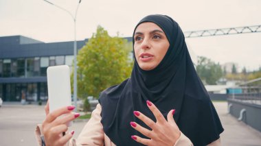 Başörtülü genç Müslüman kadın şehir sokağında video çağrısı yaparken el kol hareketi yapıyor. 