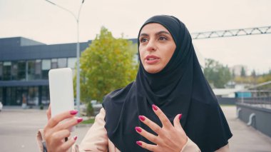 Young muslim woman in hijab gesturing while having video call on urban street  clipart