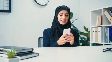 Mutlu Müslüman iş kadını masanın yanındaki defterlerin yanında akıllı telefon kullanıyor. 