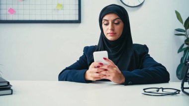 Muslim businesswoman using smartphone near notebooks on table  clipart