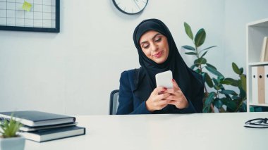 Gülümseyen Müslüman iş kadını masanın yanındaki defterlerin yanında akıllı telefon kullanıyor. 