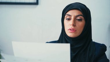 Muslim businesswoman in hijab looking at blurred documents in office  clipart