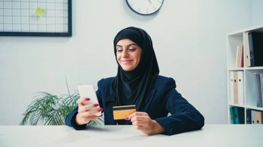 happy muslim businesswoman using smartphone and credit card in office clipart