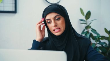 confused muslim woman in hijab looking at blurred laptop clipart