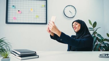 Müslüman iş kadını dudaklarını büzüyor ve ofisteki akıllı telefondan selfie çekiyor. 