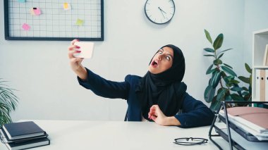 Muslim businesswoman grimacing while taking selfie on smartphone in office  clipart