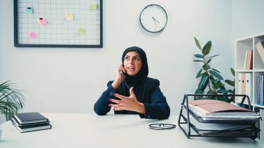 Muslim businesswoman sitting at desk and talking on smartphone in office  clipart