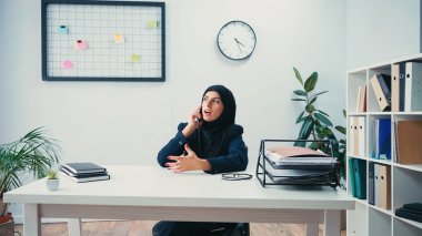 Muslim businesswoman in hijab sitting at desk and talking on smartphone in office  clipart