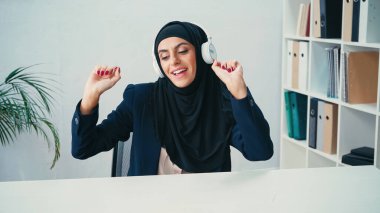 Mutlu Müslüman iş kadını tesettürlü ve kablosuz kulaklıklı ofiste müzik dinliyor. 