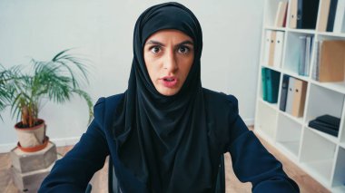 displeased arabian businesswoman in hijab looking at camera in office  clipart