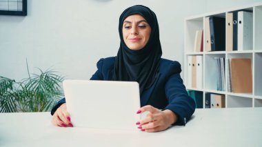 cheerful arabian woman in hijab looking at digital tablet in office  clipart