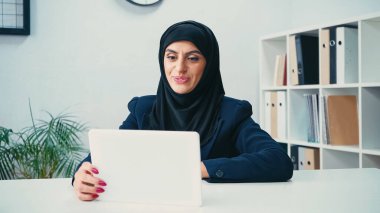smiling muslim woman in hijab looking at digital tablet in office  clipart