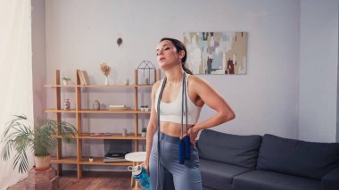 Atlama ipi ve spor şişesiyle oturma odasında duran yorgun sporcu kadın. 