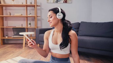 Evde akıllı telefon kullanarak gülümseyen kulaklıklı bir sporcu kadın. 