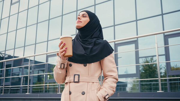 low angle view of muslim woman in hijab and trench coat holding paper cup and posing with hand in pocket near building outside 
