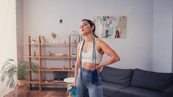 Tired sportswoman with jump rope and sports bottle standing in living room