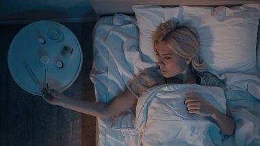 Yatak odasındaki komodinin üzerindeki termometreye bakan hasta kadının üst görüntüsü. 