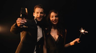 cheerful man holding glass of red wine and woman with sparkler looking at camera on black  clipart