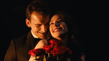 cheerful woman receiving red roses from man standing behind isolated on black  clipart