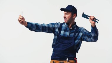 Boru anahtarı olan tamirci, beyazda izole edilmiş akıllı telefondan selfie çekiyor.