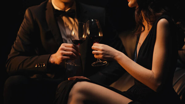 cropped view of couple clinking glasses of red wine while sitting on sofa isolated on black