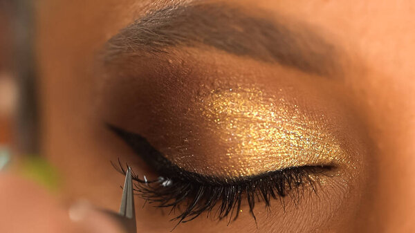 cropped view of african american woman with closed eye apply black false eyelashes with tweezers