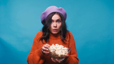 thrilled woman holding bowl with popcorn while watching film isolated on blue clipart