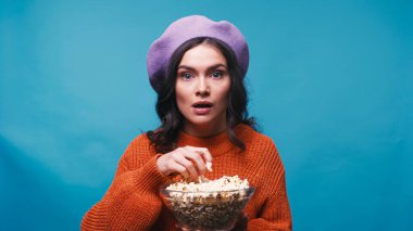 impressed woman holding bowl of popcorn while watching interesting film isolated on blue clipart