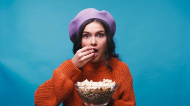 astonished woman in beret watching movie and eating popcorn isolated on blue clipart