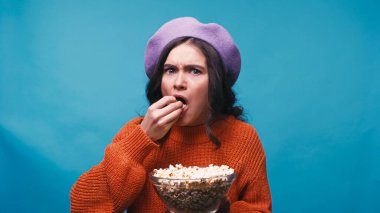 worried woman in lilac beret eating popcorn while watching movie isolated on blue clipart