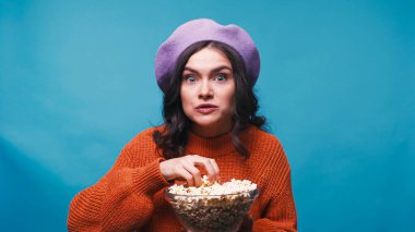 excited woman in sweater and beret holding popcorn while watching film isolated on blue clipart