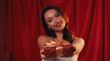 Bulanık genç bir kadın gülümsüyor ve elinde kırmızı bir hediye kutusu var.