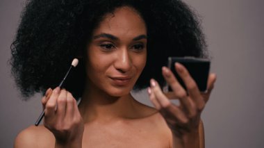 Kozmetik fırçalı genç Afro-Amerikan kadın, gri renkte izole edilmiş aynaya bakıyor.