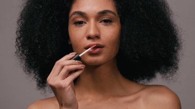 Kıvırcık Afrikalı Amerikalı kadın dudak parlatıcısı sürüyor ve gri renkli kameraya bakıyor.