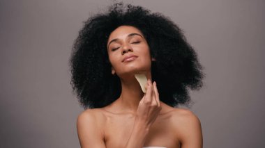 Gri üzerinde Gua sha taşı kullanan çıplak omuzlu Afrikalı Amerikalı kadın.