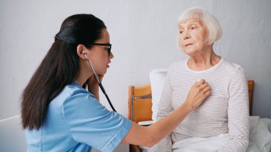 Gözlüklü bulanık hemşire yatakta yaşlı kadını muayene ediyor.