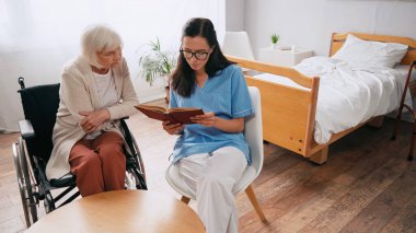 Tekerlekli sandalyedeki yaşlı kadına kitap okuyan genç bir moruk.